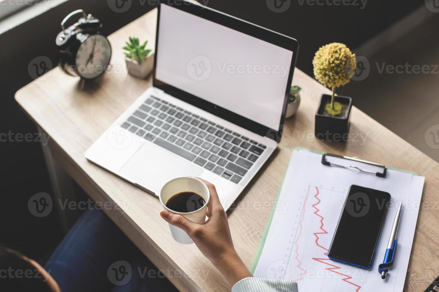 femme d'affaires assise au bureau devant un ordinateur portable à écran blanc tout en tenant une tasse de café. écran de maquette pour les annonces. concept publicitaire sur un écran. espace de copie vide sur le moniteur pour la publicité photo
