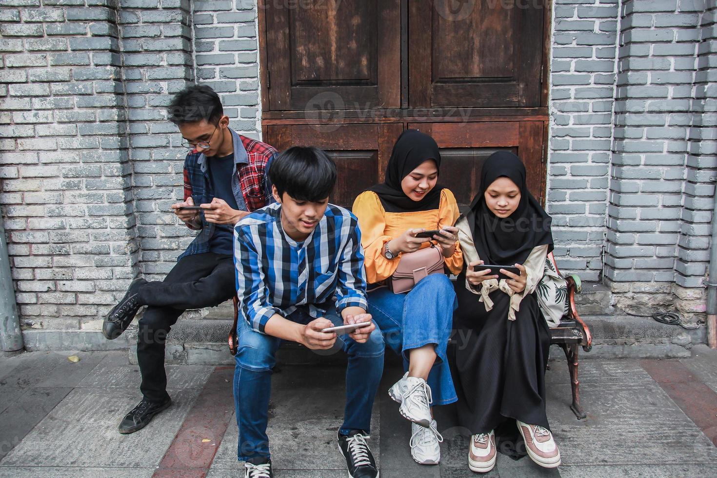 quatre jeunes amis assis à l'extérieur et regardant un téléphone portable. groupe de personnes assises sur un banc et jouant à des jeux en ligne sur un smartphone photo