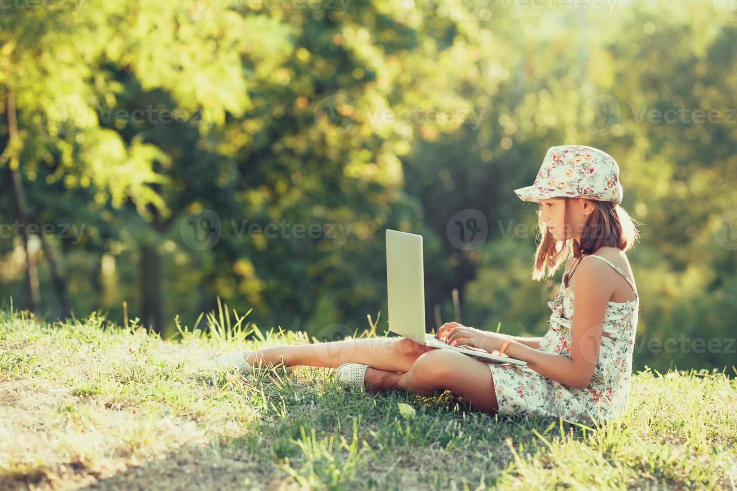 petite fille parle sur un ordinateur portable assis sur l'herbe au soleil. vêtu d'un sarafan et d'un chapeau photo