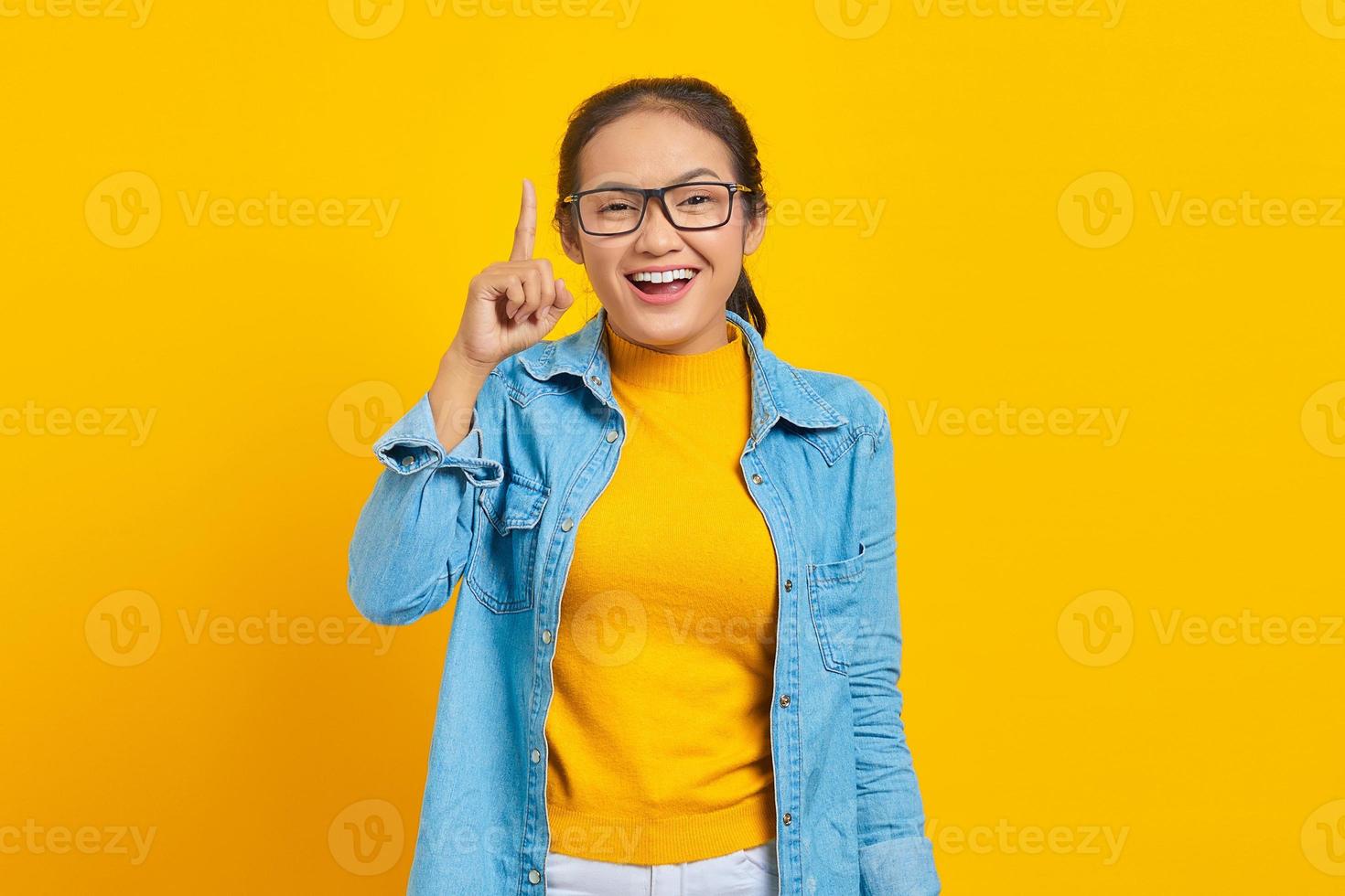 portrait d'une jeune étudiante asiatique joyeuse en vêtements denim ayant une idée créative et pointant le doigt vers le haut isolé sur fond jaune. éducation au concept d'université collégiale photo