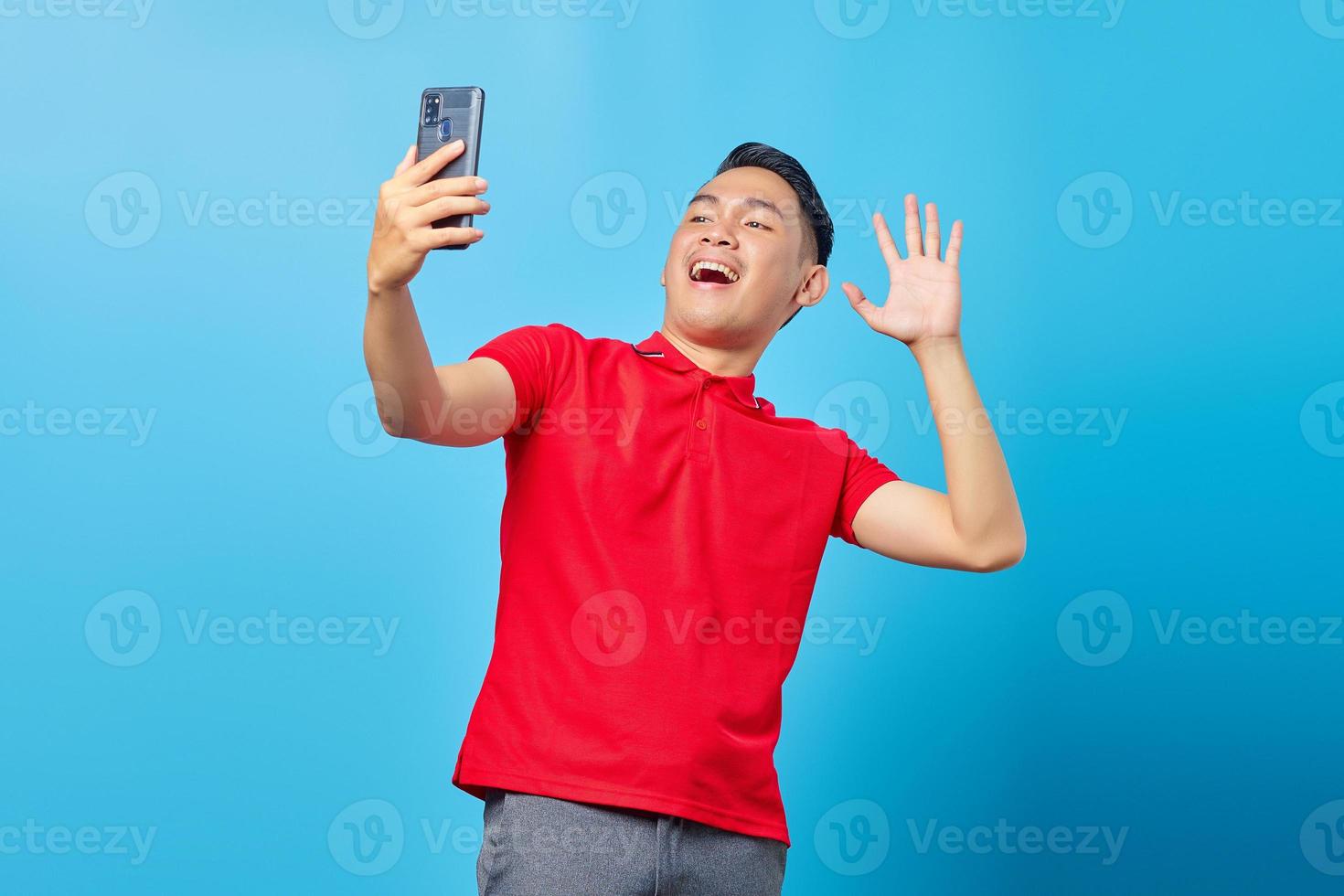 portrait d'un jeune homme asiatique joyeux faisant un appel vidéo et agitant la caméra du smartphone isolé sur fond bleu photo