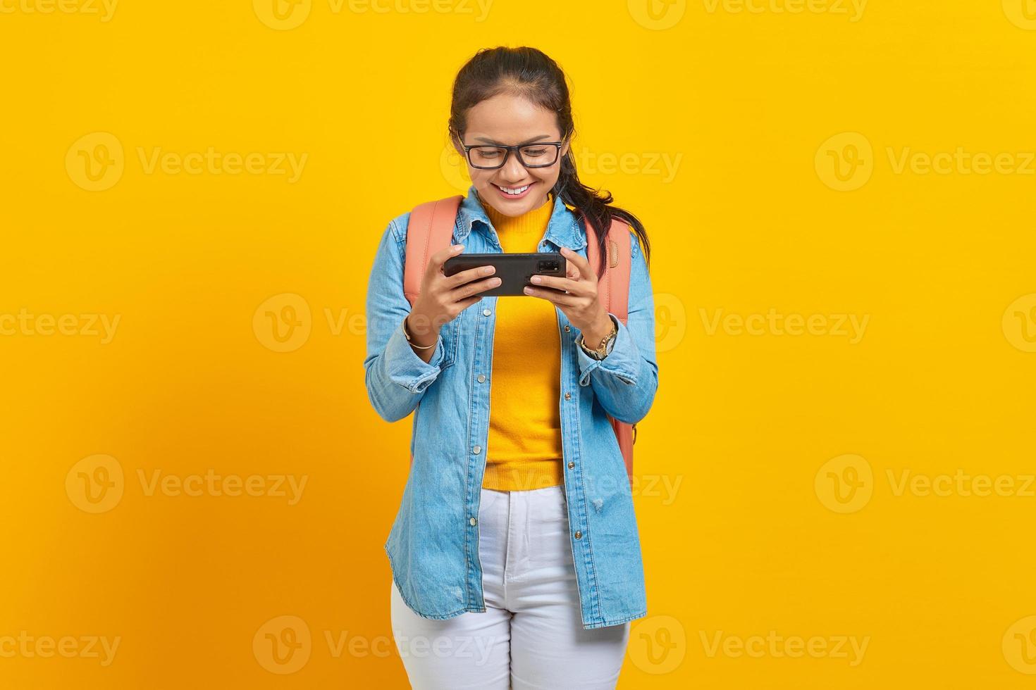 portrait d'une jeune étudiante asiatique joyeuse dans des vêtements décontractés avec sac à dos jouant au jeu vidéo sur téléphone mobile isolé sur fond jaune. éducation au concept d'université collégiale photo