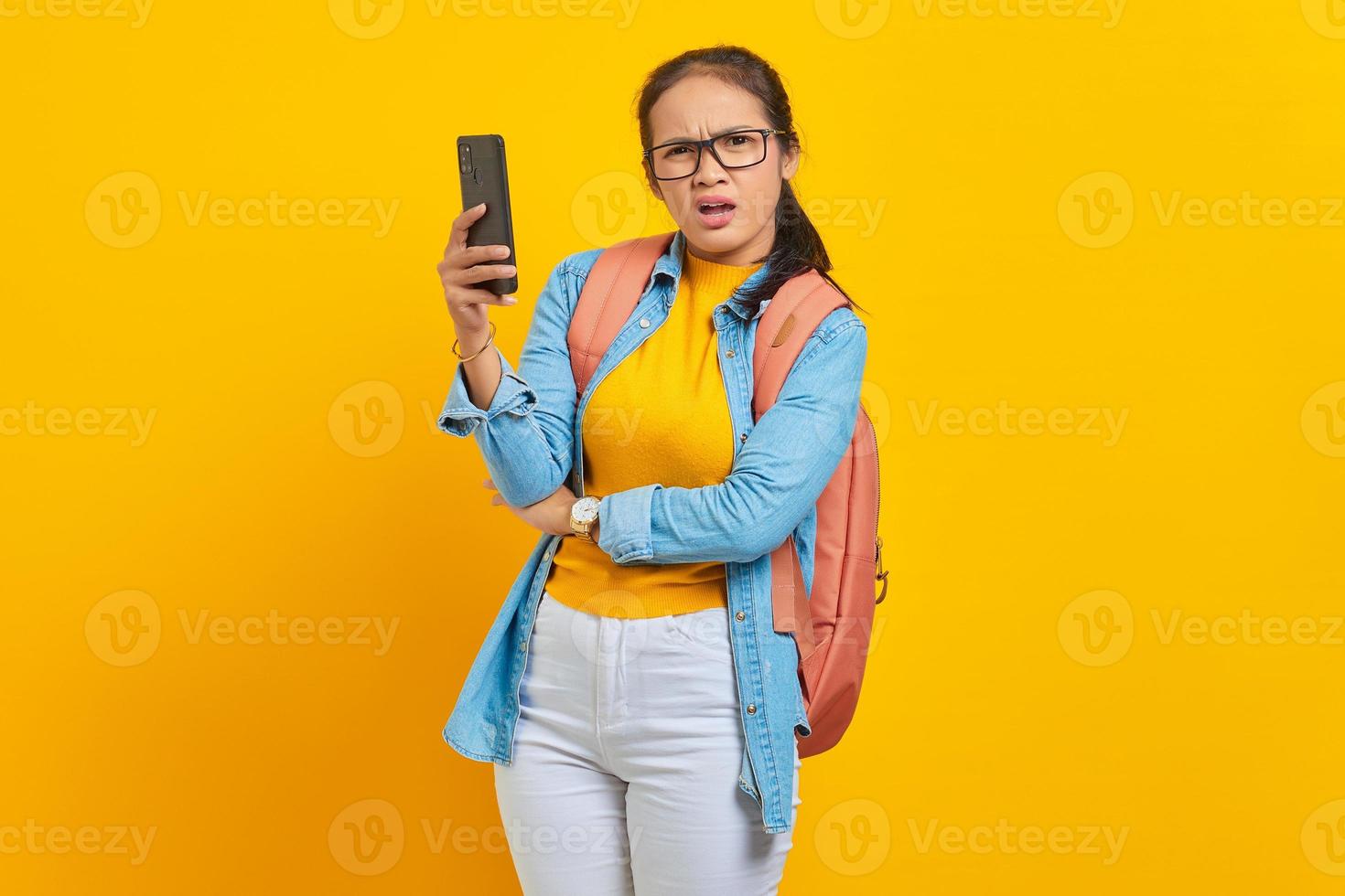 portrait d'une jeune étudiante asiatique confuse dans des vêtements décontractés avec sac à dos tenant un smartphone et regardant la caméra isolée sur fond jaune. éducation au concept d'université collégiale photo