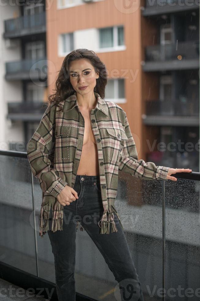 magnifique femme brune glamour avec une veste à carreaux posant sur un balcon moderne avec une vue imprenable sur la ville. portrait d'une femme élégante à la mode avec de longues jambes, un jean noir portant un balcon photo