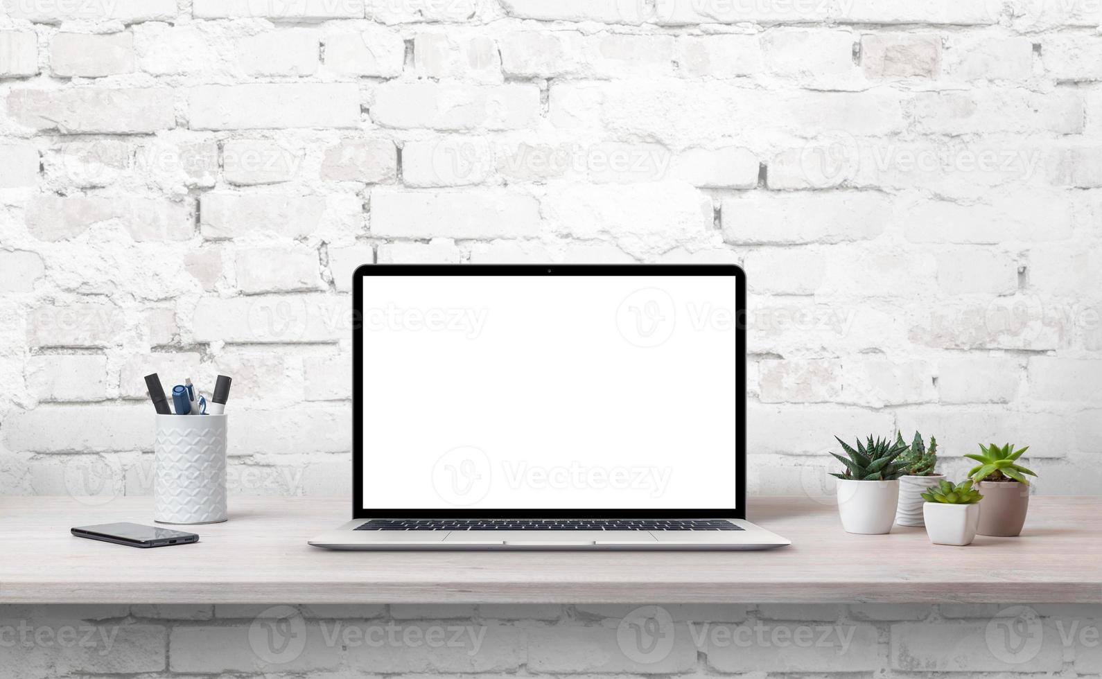 ordinateur portable sur le bureau avec écran isoalté pour maquette. plantes, téléphone et stylos à côté. mur de briques blanches en arrière-plan photo