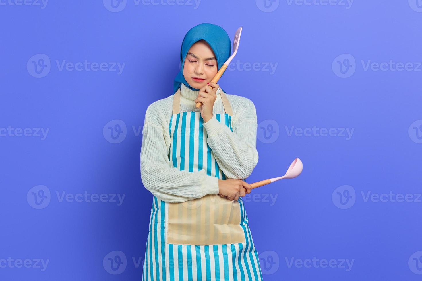 pensive jeune femme asiatique femme au foyer en tablier blanc et pull tenant une cuillère à soupe et une spatule tout en faisant le ménage isolé sur fond violet portrait en studio. notion de ménage photo