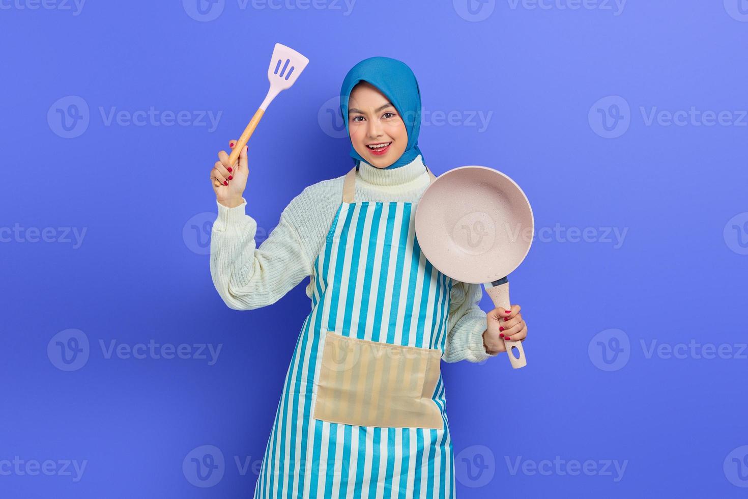 joyeuse belle femme asiatique portant un tablier tenant une spatule et une poêle à frire tout en faisant des tâches ménagères isolées sur fond violet. notion de ménage photo