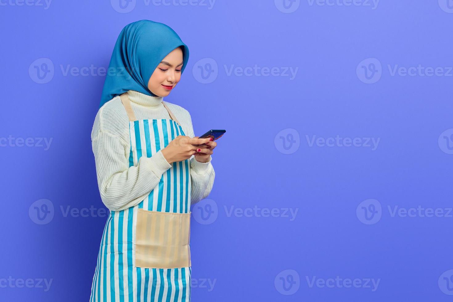 portrait d'une jeune femme musulmane asiatique joyeuse dans les années 20 portant le hijab et un tablier tapant des messages sms dans un téléphone portable isolé sur fond violet. concept de mode de vie musulman femme au foyer photo