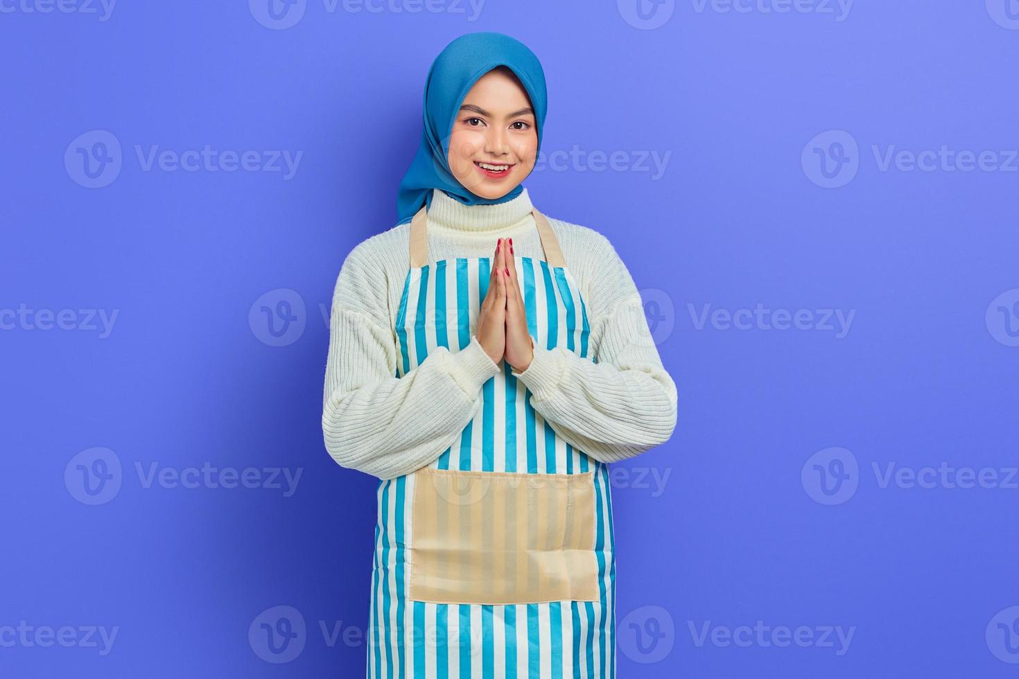 joyeuse jeune femme musulmane asiatique portant le hijab et le tablier, signe ouvert pour saluer et saluer les clients isolés sur fond violet. concept de mode de vie musulman femme au foyer photo