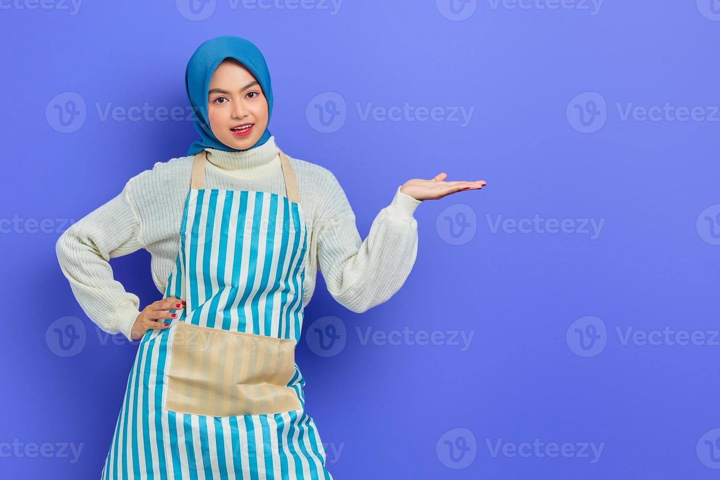 portrait d'une jeune femme musulmane asiatique souriante en hijab et tablier rayé, pointant de côté avec les mains tout en faisant le ménage isolé sur fond violet. concept de mode de vie musulman femme au foyer photo