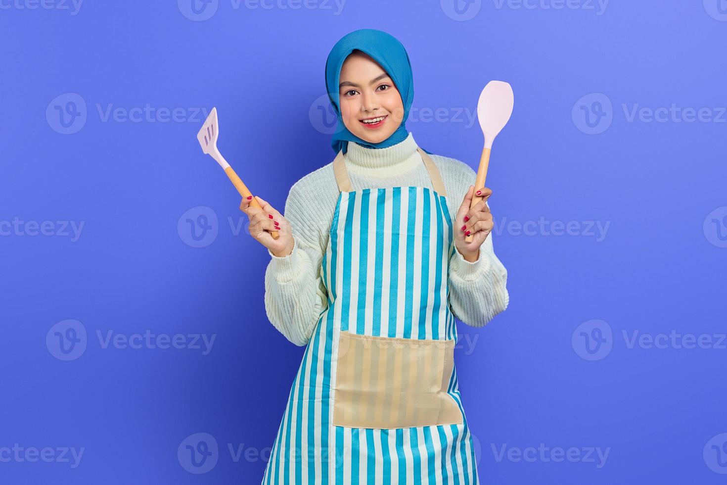 joyeuse belle femme asiatique femme au foyer en tablier tenant une soupe à la spatule et à la cuillère tout en faisant le ménage isolé sur fond violet. notion de ménage photo