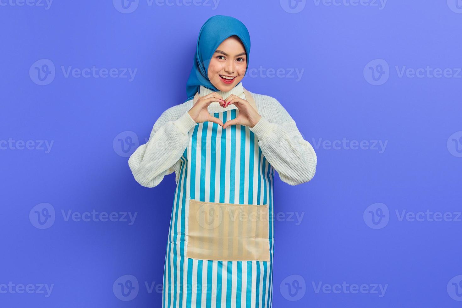 portrait d'une jeune femme au foyer souriante en hijab et tablier rayé, montrant le coeur en forme avec les mains isolées sur fond violet. concept de mode de vie musulman femme au foyer photo