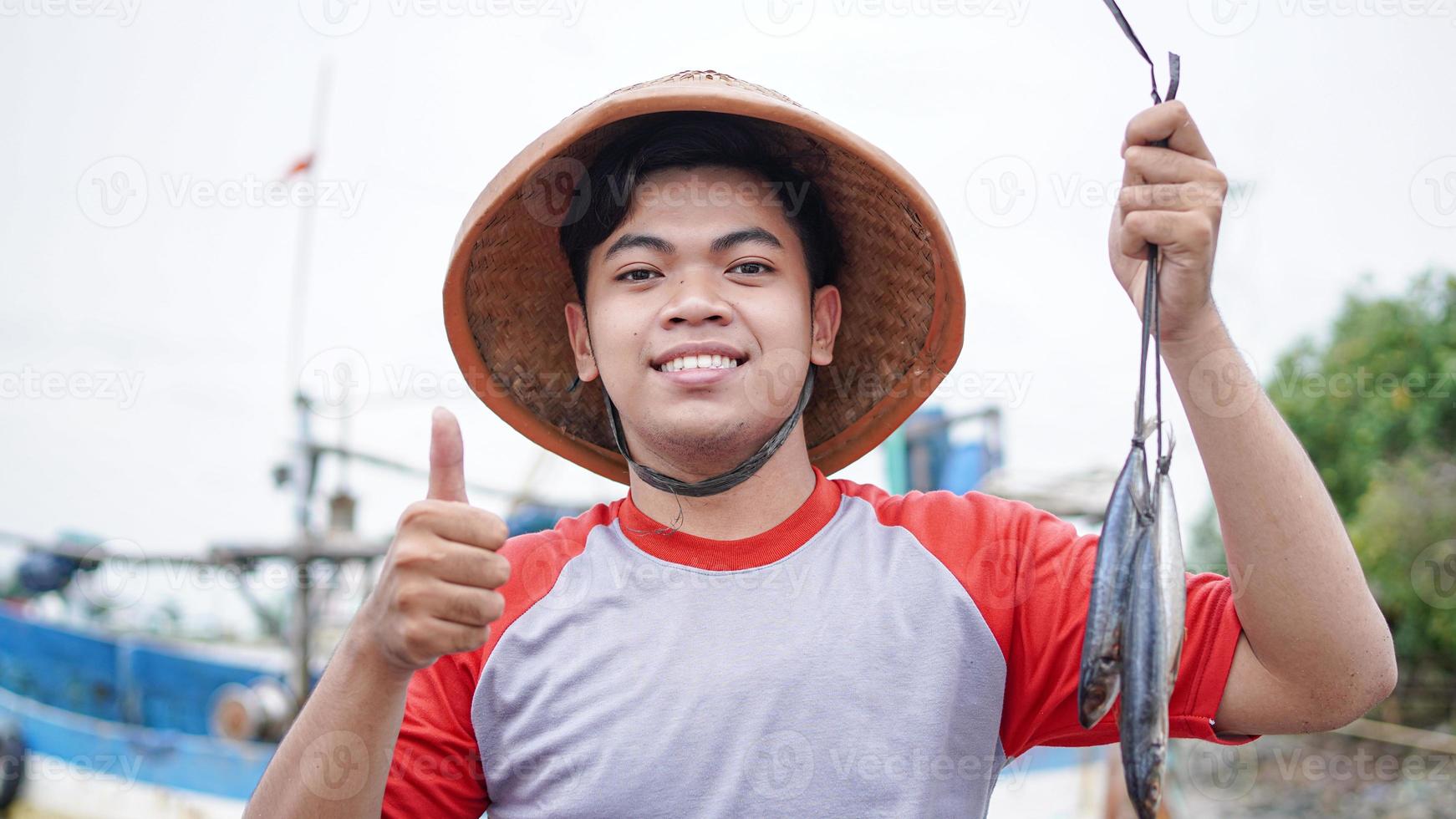 heureux jeune pêcheur sur la plage tenant son poisson et montre devant son bateau photo