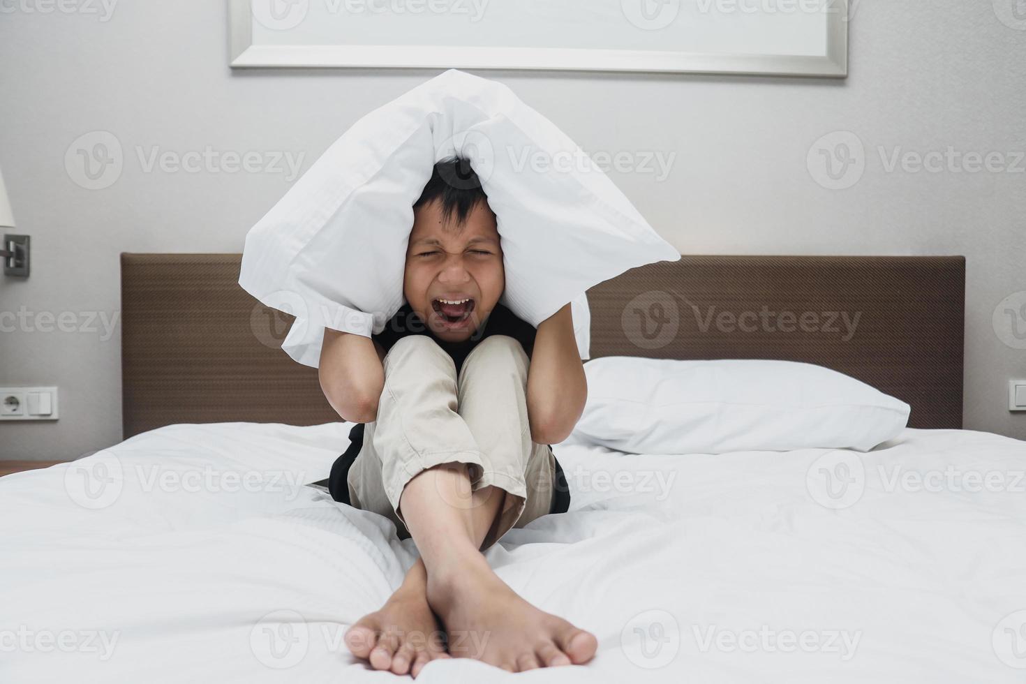triste garçon asiatique couvrant ses oreilles par un oreiller en criant sur le lit photo