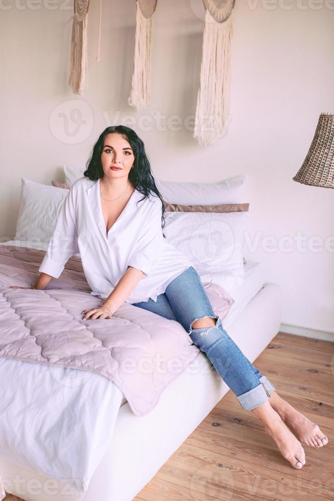 portrait d'une belle femme aux cheveux noirs en chemise blanche et jeans assis triste sur son lit. émotions, crise, maison, concept d'intérieur photo