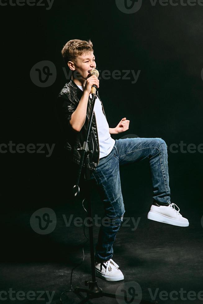 portrait d'adolescent caucasien en t-shirt blanc, jean bleu et veste en cuir avec microphone chantant sur fond sombre. concept de passe-temps et de gloire photo