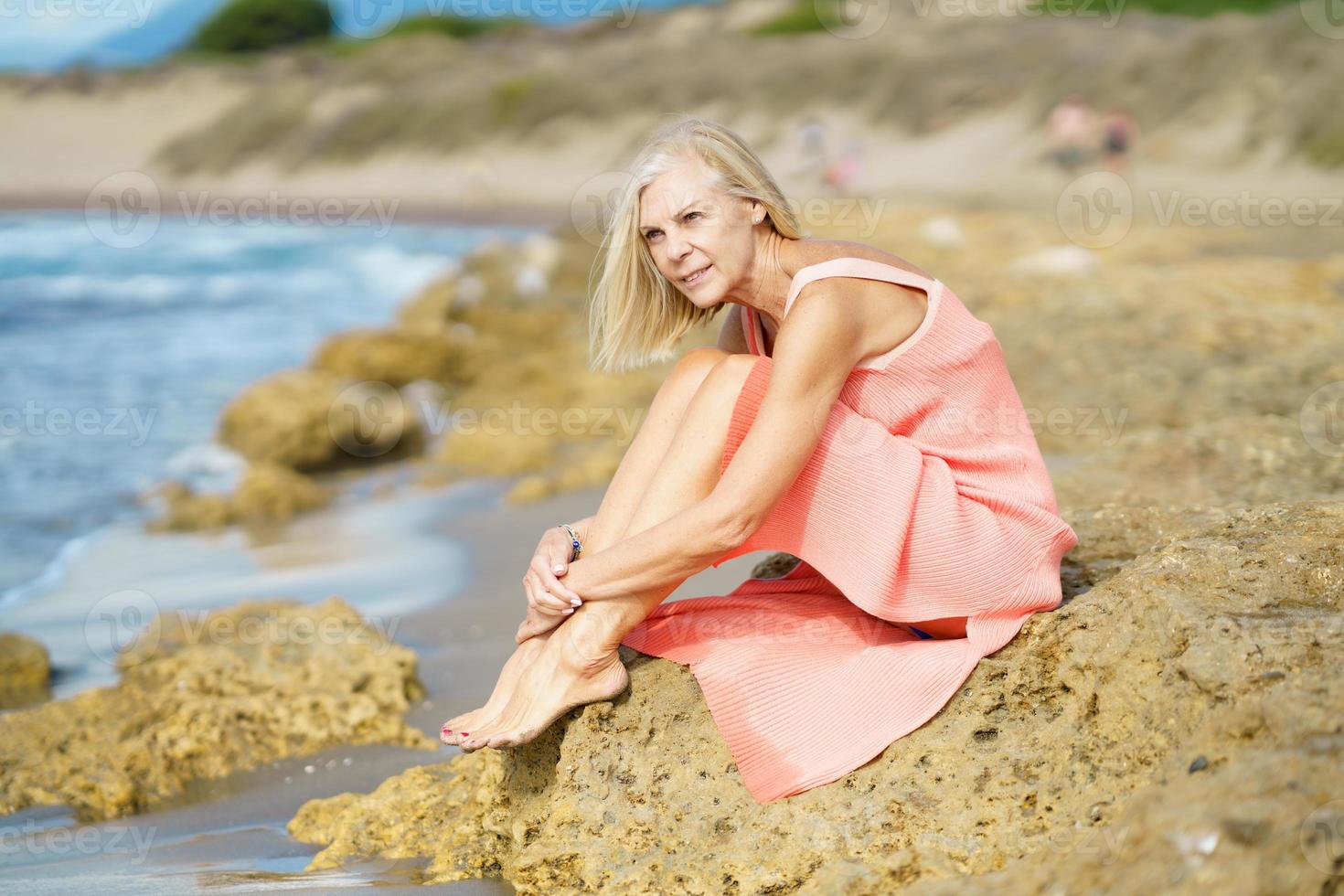 femme mûre assise sur des rochers au bord d'une plage tropicale, vêtue d'une belle robe orange. photo