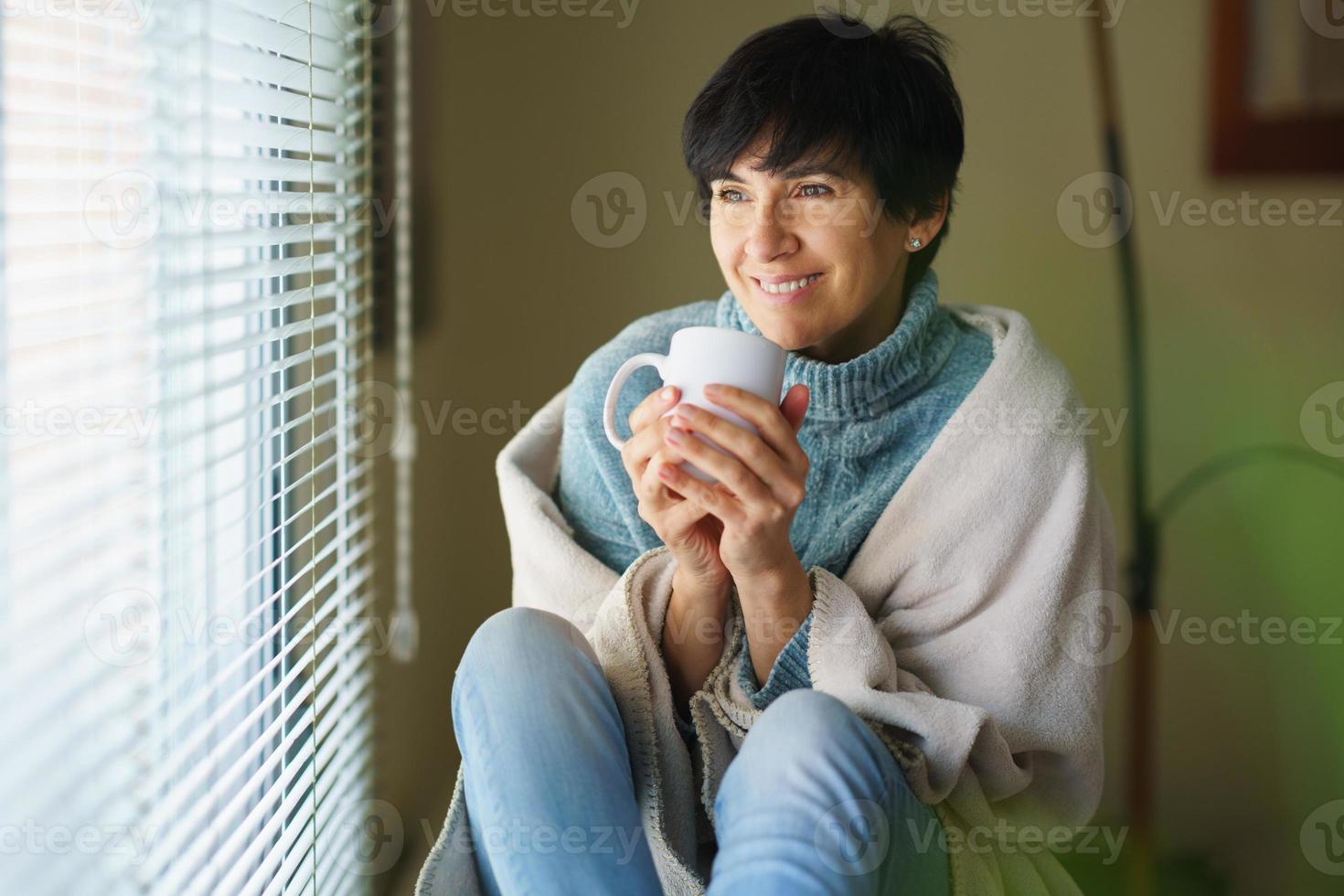 heureuse femme d'âge moyen souriant près d'une fenêtre, buvant une tasse de café. photo