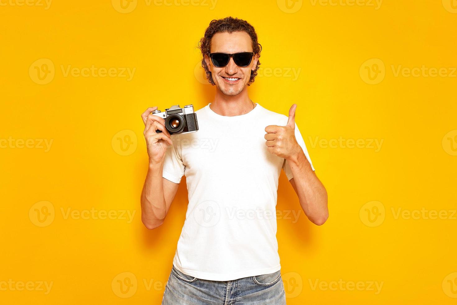 photographe masculin avec appareil photo rétro à la main donne un coup de pouce vêtu de vêtements décontractés, t-shirt blanc, jeans, lunettes de soleil isolés sur un mur de studio jaune. concept - personnes, créativité, technologie