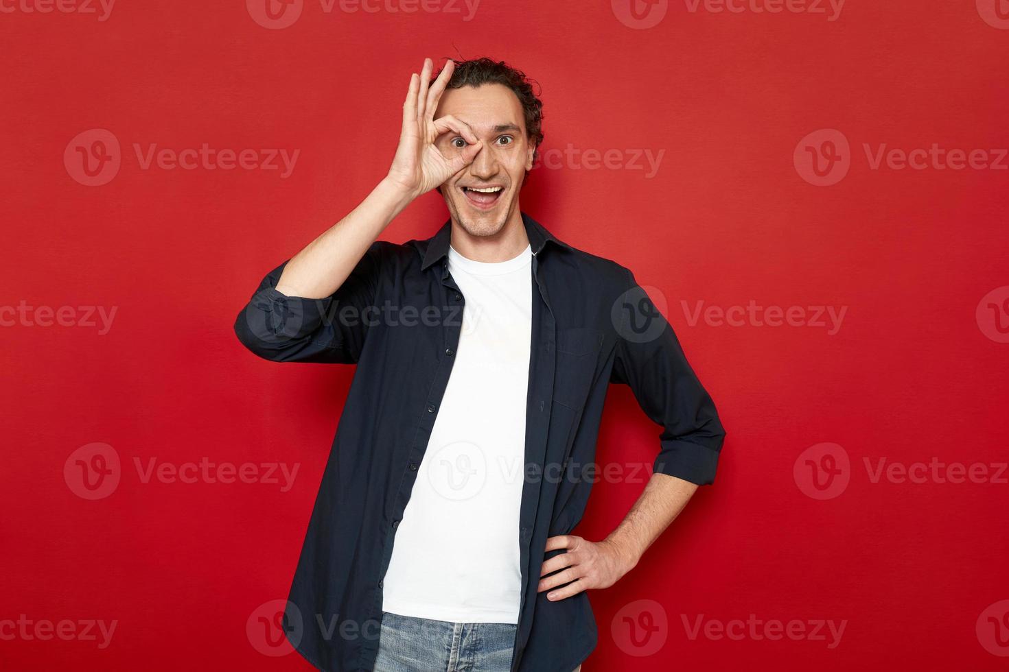 l'homme agité apporte sa main à son œil montrant le signe ok. vêtus de vêtements décontractés. isolé sur fond rouge avec un espace pour le texte publicitaire. concept - personnes, langage corporel, chance, victoire, planification photo