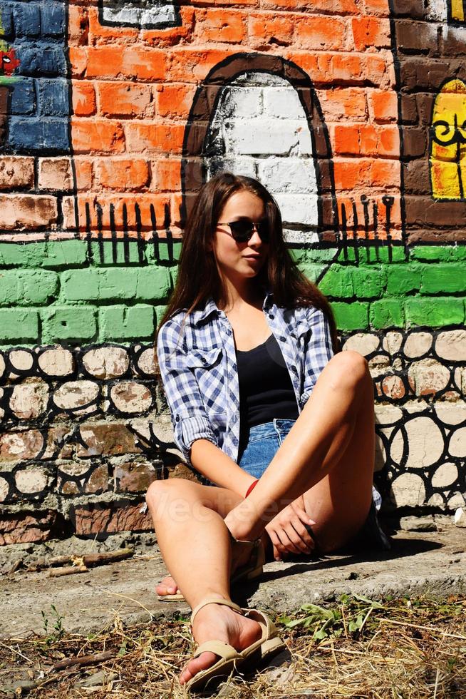 portrait d'une belle adolescente en lunettes de soleil portées sur une chemise à carreaux et un short en jean, contre un mur avec un élément de graffiti. photo