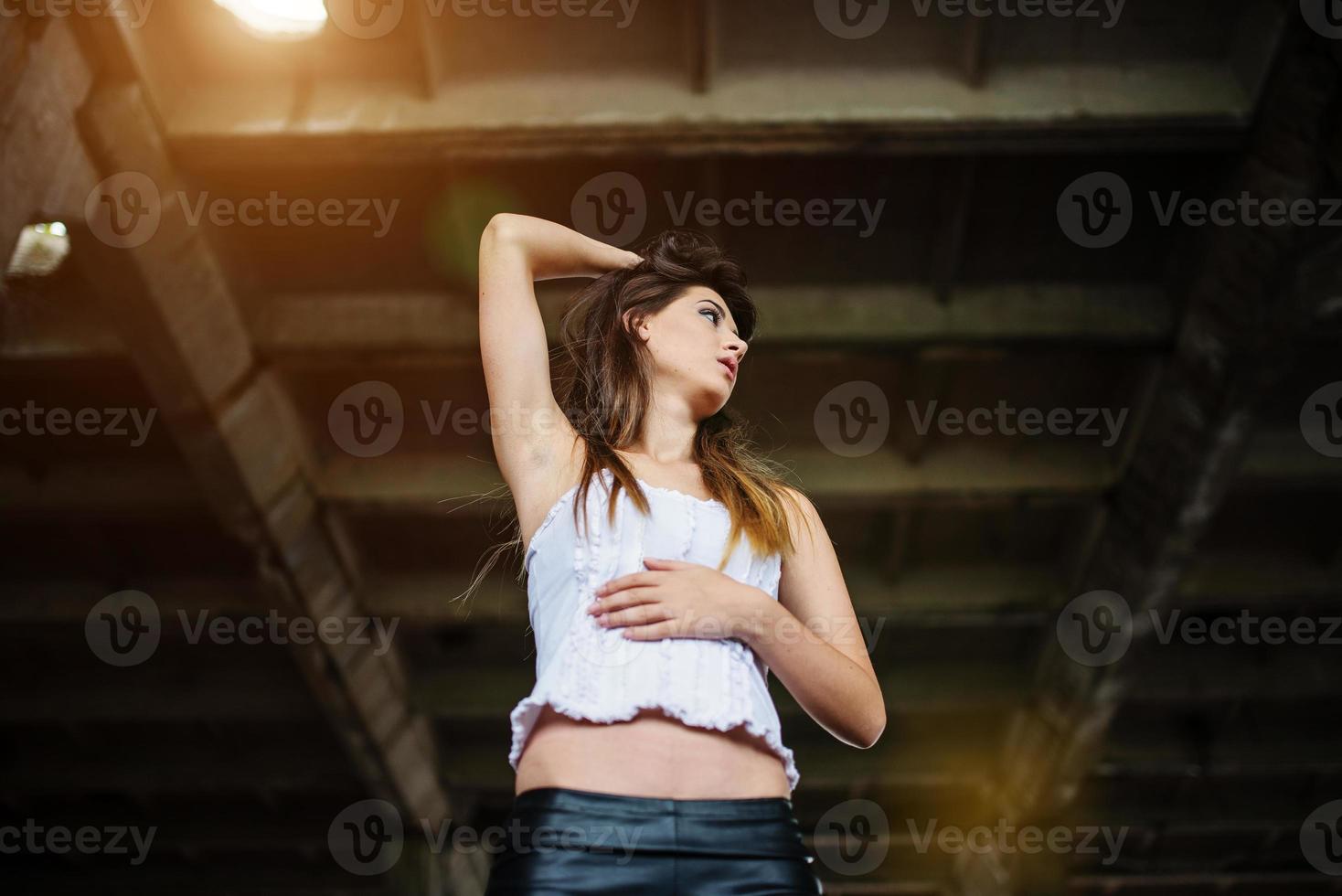 portrait d'une jeune fille brune mignonne portant un pantalon en cuir noir et un chemisier blanc posé sur un lieu abandonné. photo