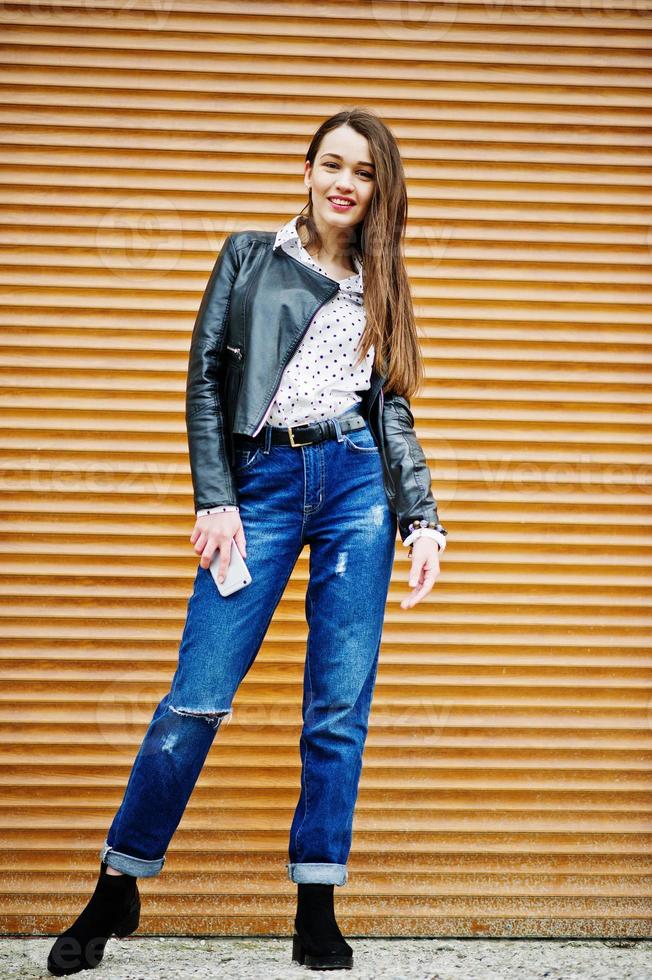 portrait d'une jeune fille élégante portant une veste en cuir et un jean déchiré avec un téléphone portable à la main texture d'obturation d'arrière-plan. style de modèle de mode de rue. photo