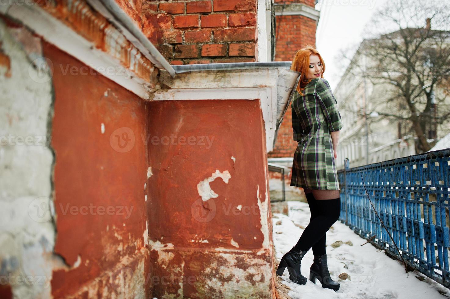 un portrait en plein air d'une jeune jolie fille aux cheveux rouges portant une robe à carreaux avec des sacs à dos girly debout sur le fond du mur de briques en journée d'hiver. photo