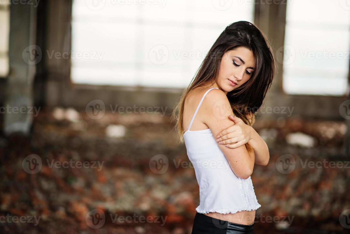 portrait d'une jeune fille brune mignonne portant un pantalon en cuir noir et un chemisier blanc posé sur un lieu abandonné. photo