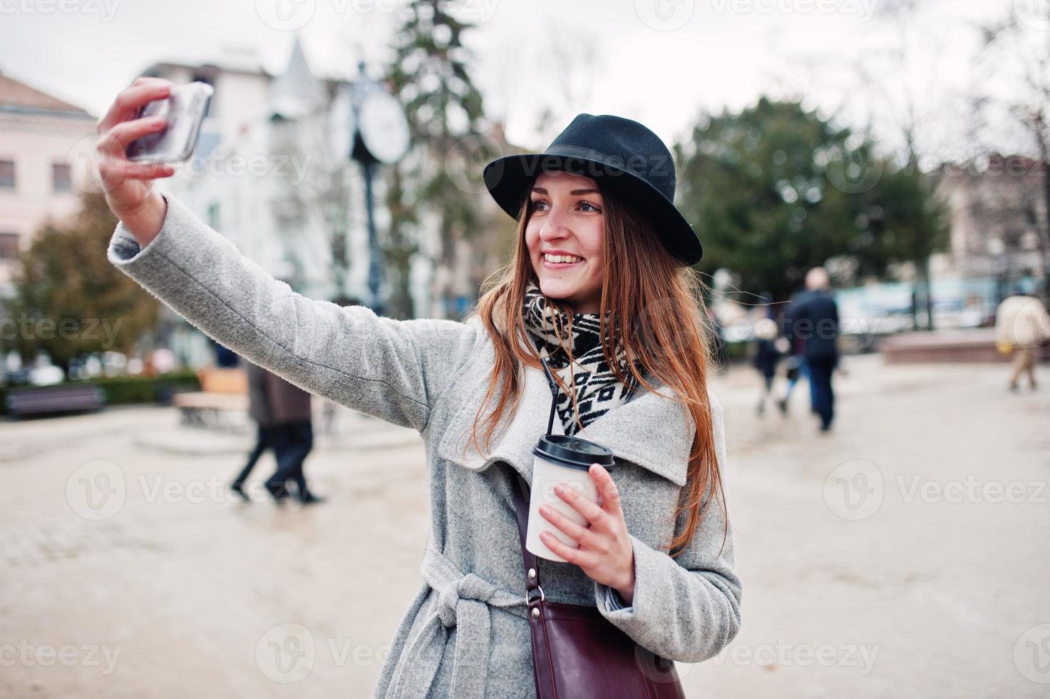 jeune fille modèle dans un manteau gris et un chapeau noir avec un sac à main en cuir sur les épaules reste avec une tasse de café en plastique et fait du selfie dans la rue de la ville. photo