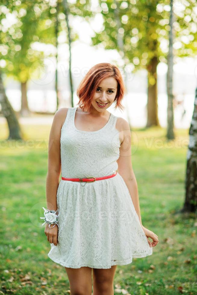 portrait d'une fille aux cheveux rouges vêtue d'une robe blanche contre le parc à la fête de poule. photo