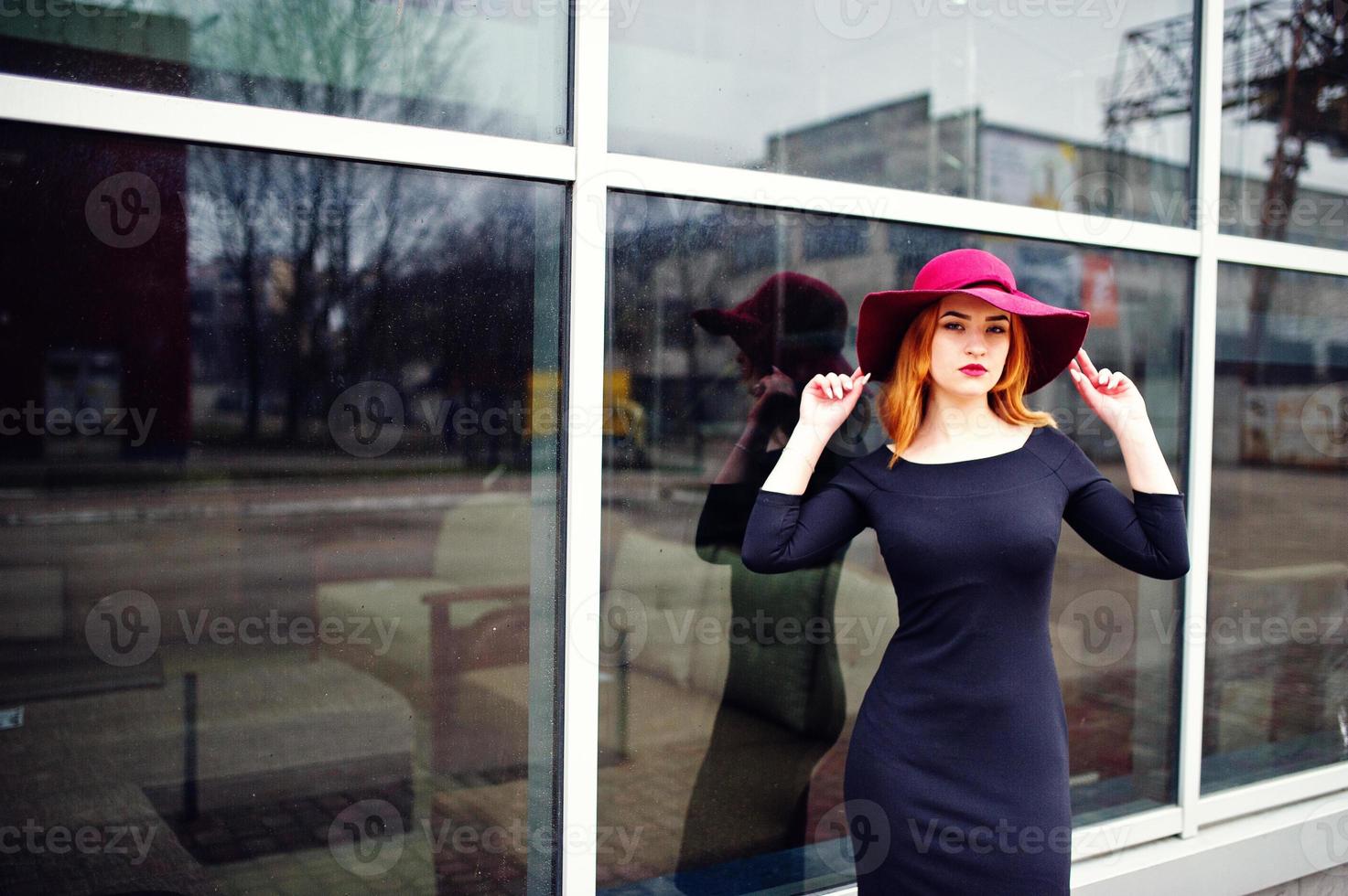 portrait d'une fille aux cheveux rouges fashion sur un chapeau rouge et une robe noire avec un maquillage lumineux posé contre une grande fenêtre. filtres instagram de style photo tonique.