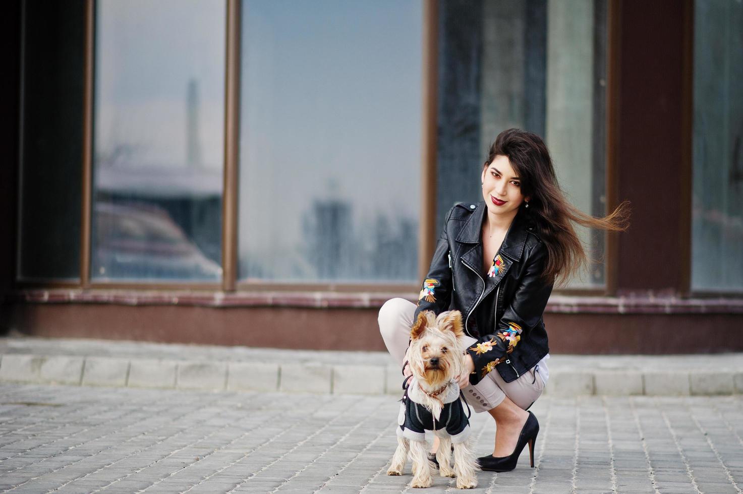 brune gitane avec chien yorkshire terrier posé contre de grandes fenêtres maison. le modèle porte une veste en cuir et un t-shirt avec ornement, un pantalon et des chaussures à talons hauts. photo