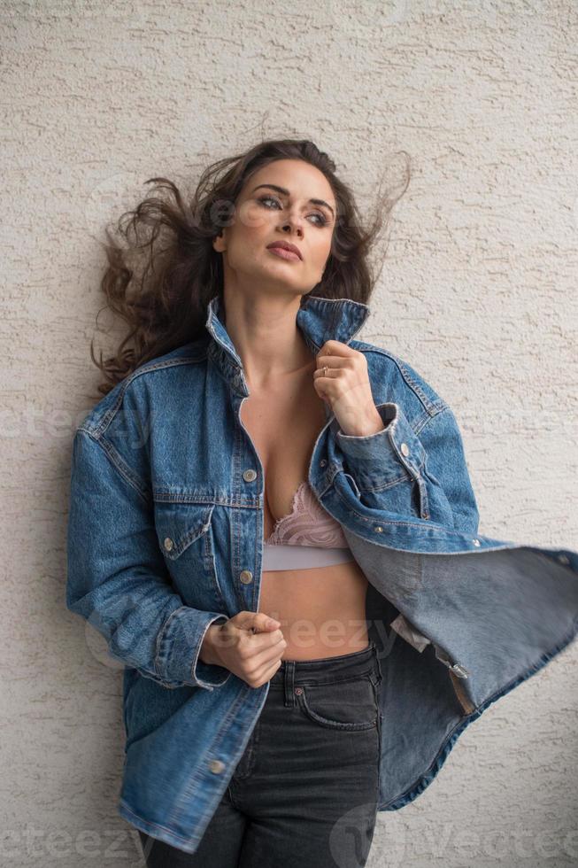 magnifique femme brune glamour avec une veste bleue posant sur un balcon moderne avec une vue imprenable sur la ville. portrait d'une femme élégante à la mode avec de longs cheveux, un jean noir portant sur un balcon photo