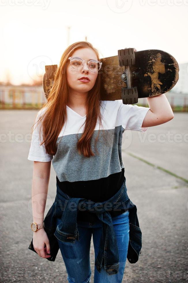 jeune adolescente urbaine avec planche à roulettes, porter des lunettes, une casquette et un jean déchiré sur le terrain de sport de la cour au coucher du soleil. photo