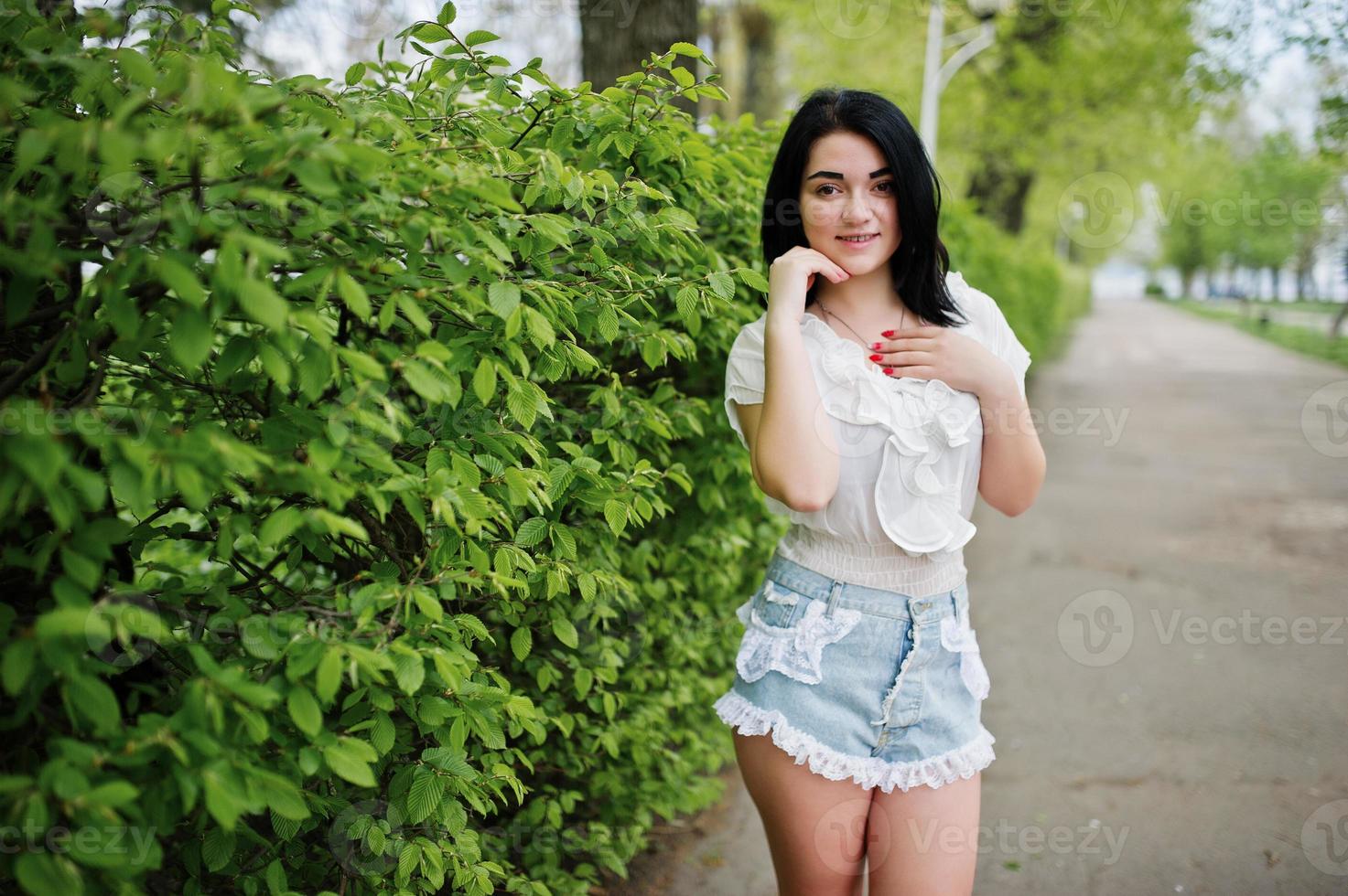 portrait d'une fille brune sexy sur un short en jean pour femme et un chemisier blanc contre des buissons de printemps verts. photo