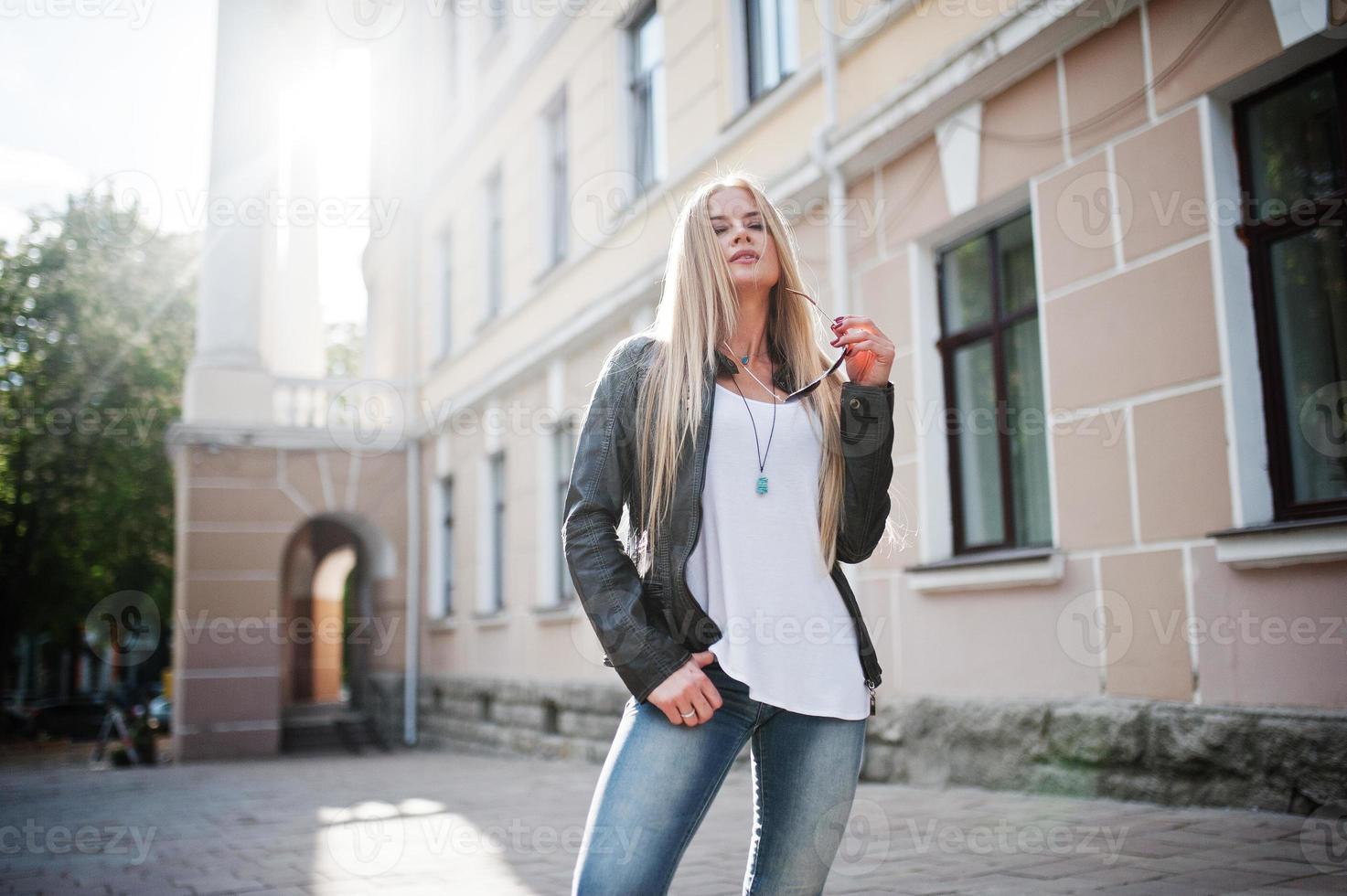femme blonde élégante portant des jeans, des lunettes de soleil et une veste posée dans la rue au soleil. portrait de modèle urbain de mode. photo
