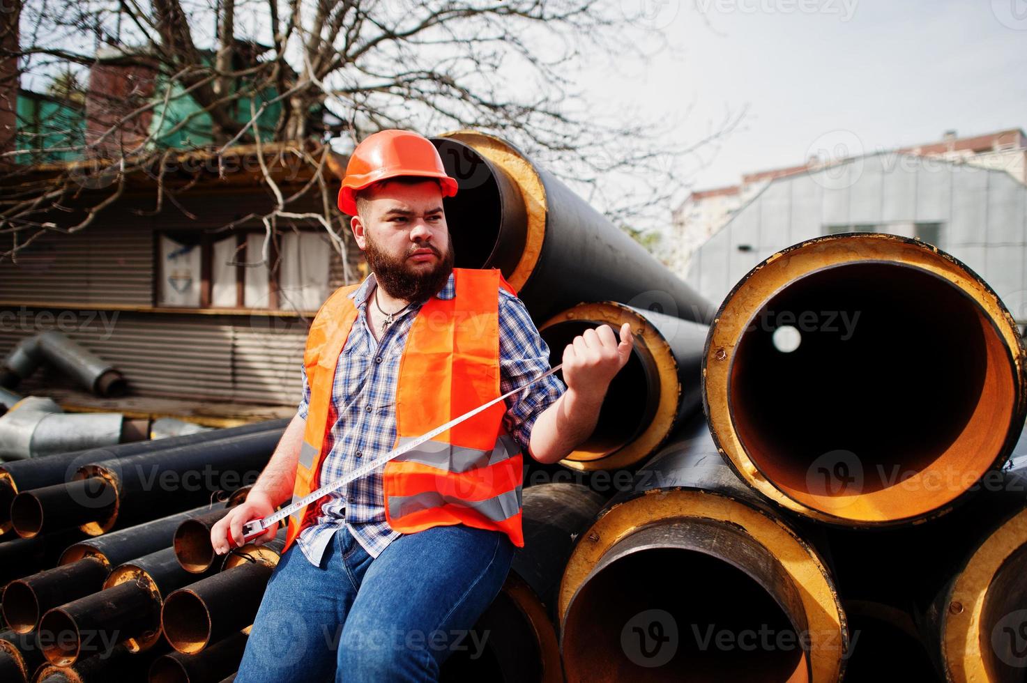 travailleur de barbe brutal homme costume travailleur de la construction dans un casque orange de sécurité près de tuyaux en acier avec un ruban à mesurer à portée de main. photo