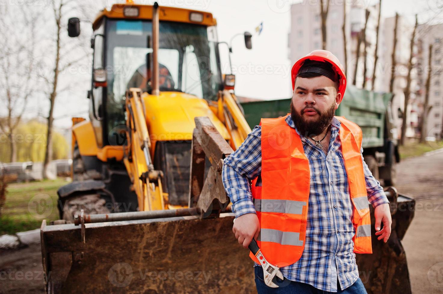 travailleur de barbe brutal homme costume travailleur de la construction dans un casque orange de sécurité, contre traktor avec une clé à molette à portée de main. photo