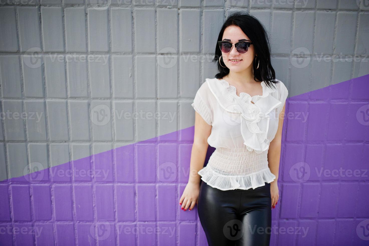 portrait d'une jeune fille brune sur un pantalon en cuir pour femmes et un chemisier blanc, des lunettes de soleil, contre un mur violet et gris. photo