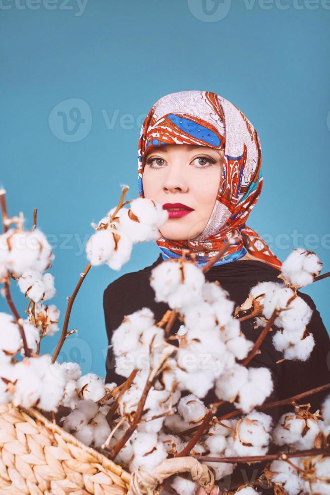 orient femme dans un mouchoir coloré tenant un panier avec des branches de coton sur fond bleu. cueilleur de coton photo