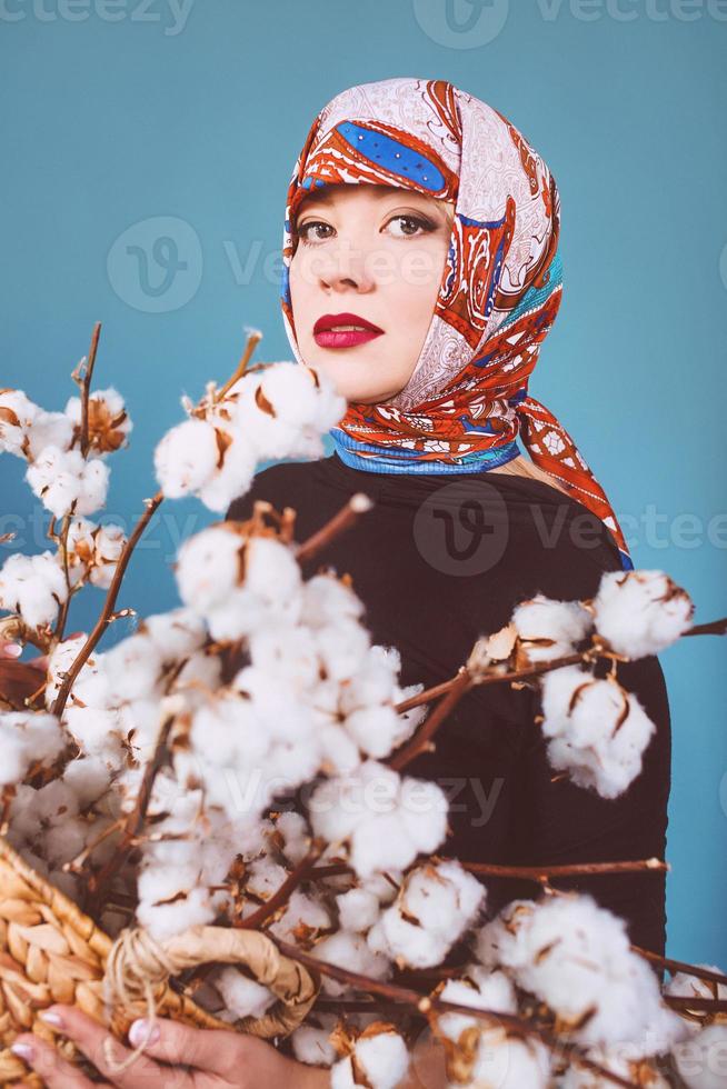 orient femme dans un mouchoir coloré tenant un panier avec des branches de coton sur fond bleu. cueilleur de coton photo