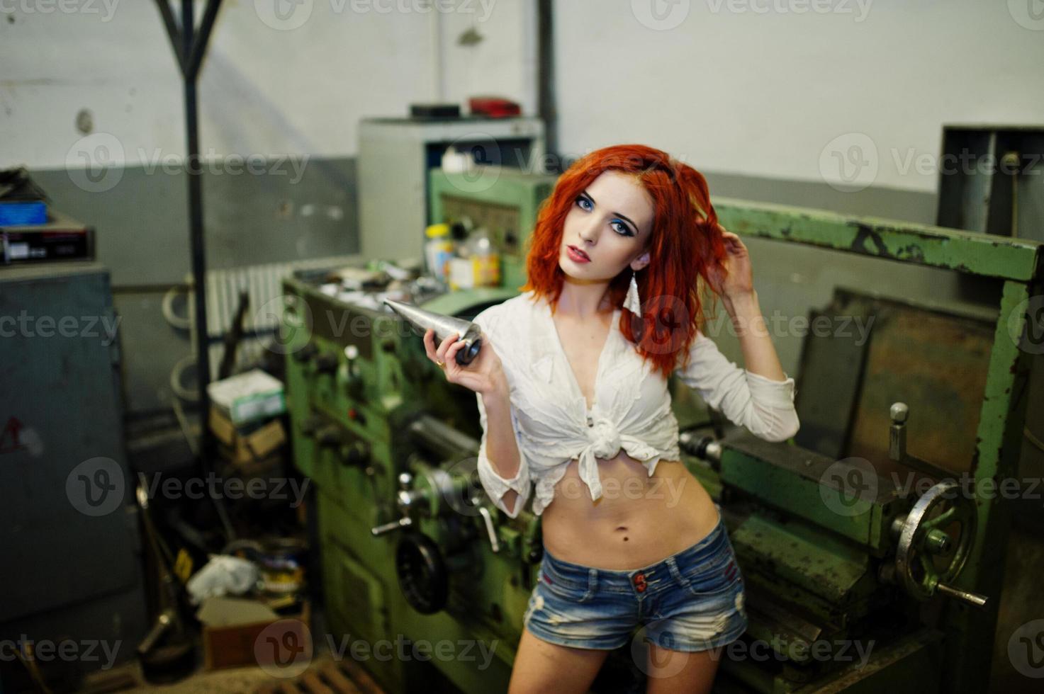 fille aux cheveux rouges portant un short en jean court et un chemisier blanc posé sur une machine industrielle à l'usine. photo