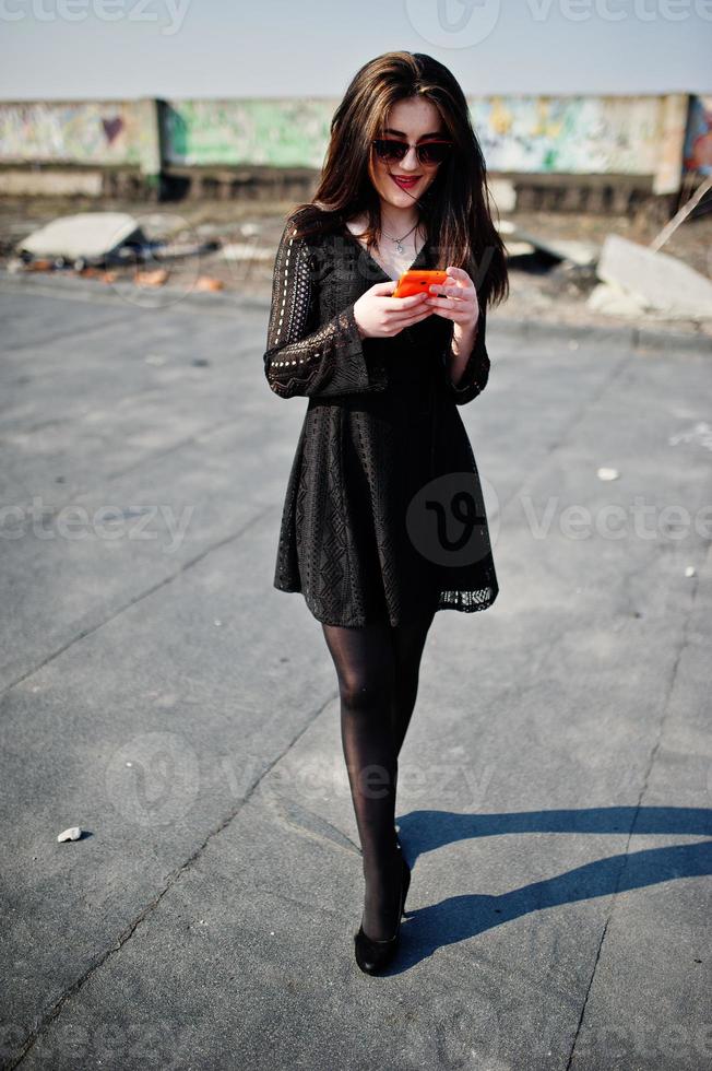 portrait fille brune aux lèvres rouges et téléphone portable orange à portée de main, vêtue d'une robe noire, lunettes de soleil posées sur le toit. mannequin de rue. photo