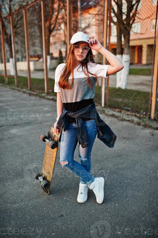 jeune adolescente urbaine avec planche à roulettes, porter des lunettes, une casquette et un jean déchiré sur le terrain de sport de la cour. photo