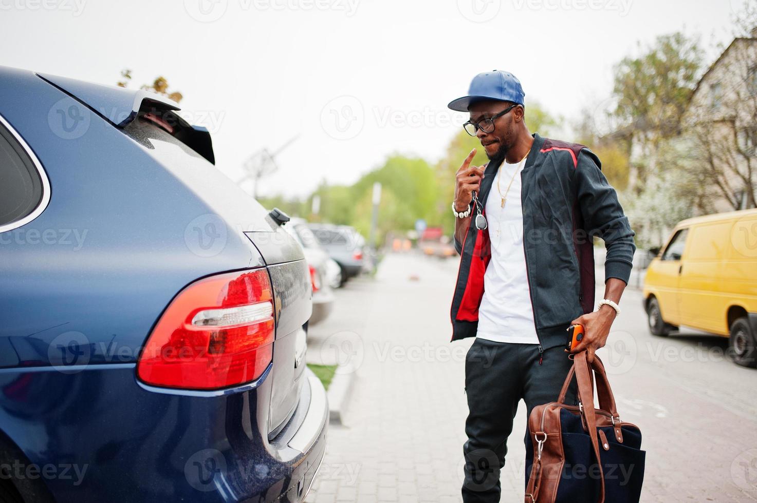 portrait d'un homme afro-américain élégant sur des vêtements de sport, une casquette et des lunettes marchant avec un sac à main et un coffre de voiture ouvert. les hommes noirs modèlent la mode de la rue. photo