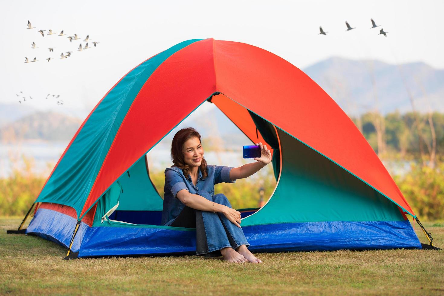 une belle femme assise dans une tente de camping et utilisant un smartphone prenant un selfie pour elle-même avec un grand lac en arrière-plan et un groupe d'oiseaux volant le matin. photo