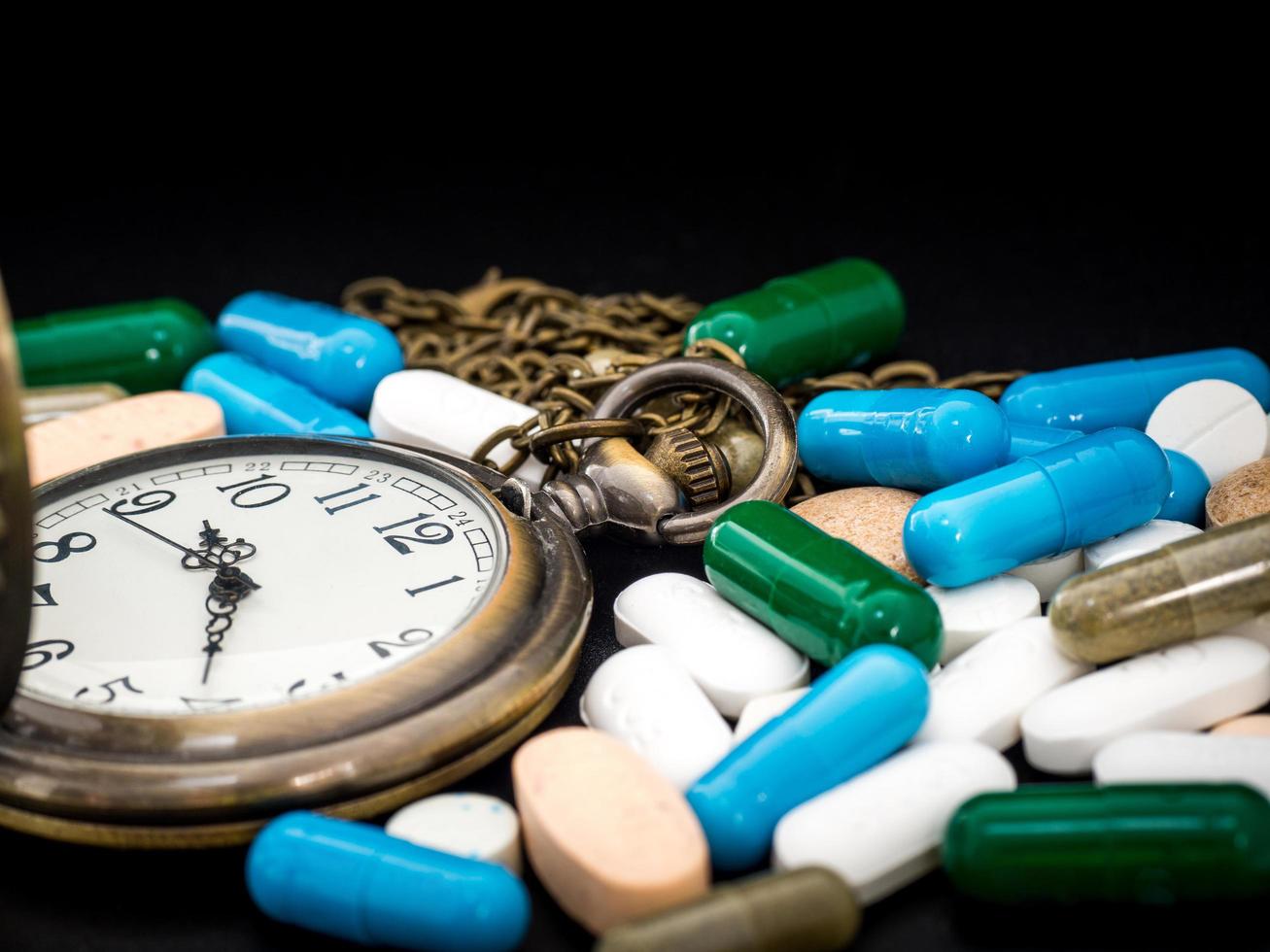 horloge antique sur multicolore de médicament et capsule est sur le fond noir. fermer. nous sommes contre les médicaments anti-médicaments, cure dans un récipient pour la santé. tas de pilules à capsules rondes blanches bleues vertes photo