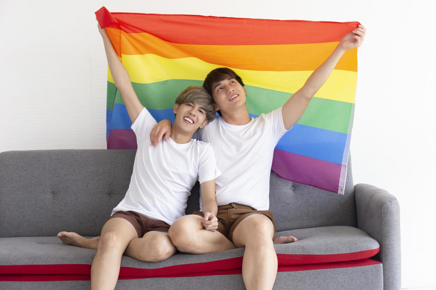 un couple masculin avec un homme asiatique assis sur un canapé, tenant un drapeau multicolore avec des signes de lgbt sur la tête, exprimant ouvertement des hommes homosexuels acceptant les idées lgbt. photo