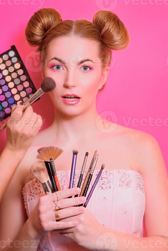 jolie femme blonde avec un maquillage coloré avec des pinceaux cosmétiques et des ombres en elle et maquille les mains du maître photo